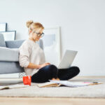 Female student sitting on floor of her apartment with laptop and notes studying